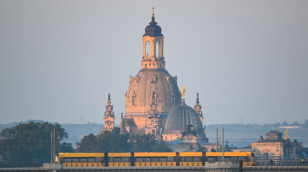 Eine Straßenbahn der Dresdner Verkehrsbetriebe (DVB) fährt am Morgen vor der Altstadtkulisse mit der Frauenkirche (l) und der Kuppel der Kunstakedmie mit dem Engel «Fama» über die Albertbrücke. / Foto: Robert Michael/dpa/Archivbild