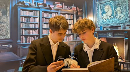 Zwei Jugendliche in schwarzen Anzügen lesen in einer Bibliothek in einem großen Buch.