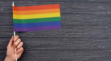 Hand hält kleine Regenbogenfahne vor dunklem Holz-Hintergrund – Symbol für queere Sichtbarkeit und Solidarität.