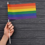 Hand hält kleine Regenbogenfahne vor dunklem Holz-Hintergrund – Symbol für queere Sichtbarkeit und Solidarität.