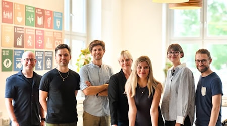 Zusammen stehen die Teammitglieder am Tisch. V.l.n.r. Michael Jenkner, Toni Koç, Matthias Damert, Silke Nyvlt, Franziska Kramer, Emma Deeken und Steve Grundig. Sie stehen gemeinsam im Co-Working Space Impact Hub Dresden, dem Firmensitz von plant values, vor den 17 SDGs.