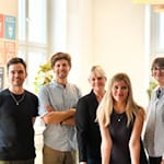 Zusammen stehen die Teammitglieder am Tisch. V.l.n.r. Michael Jenkner, Toni Koç, Matthias Damert, Silke Nyvlt, Franziska Kramer, Emma Deeken und Steve Grundig. Sie stehen gemeinsam im Co-Working Space Impact Hub Dresden, dem Firmensitz von plant values, vor den 17 SDGs.