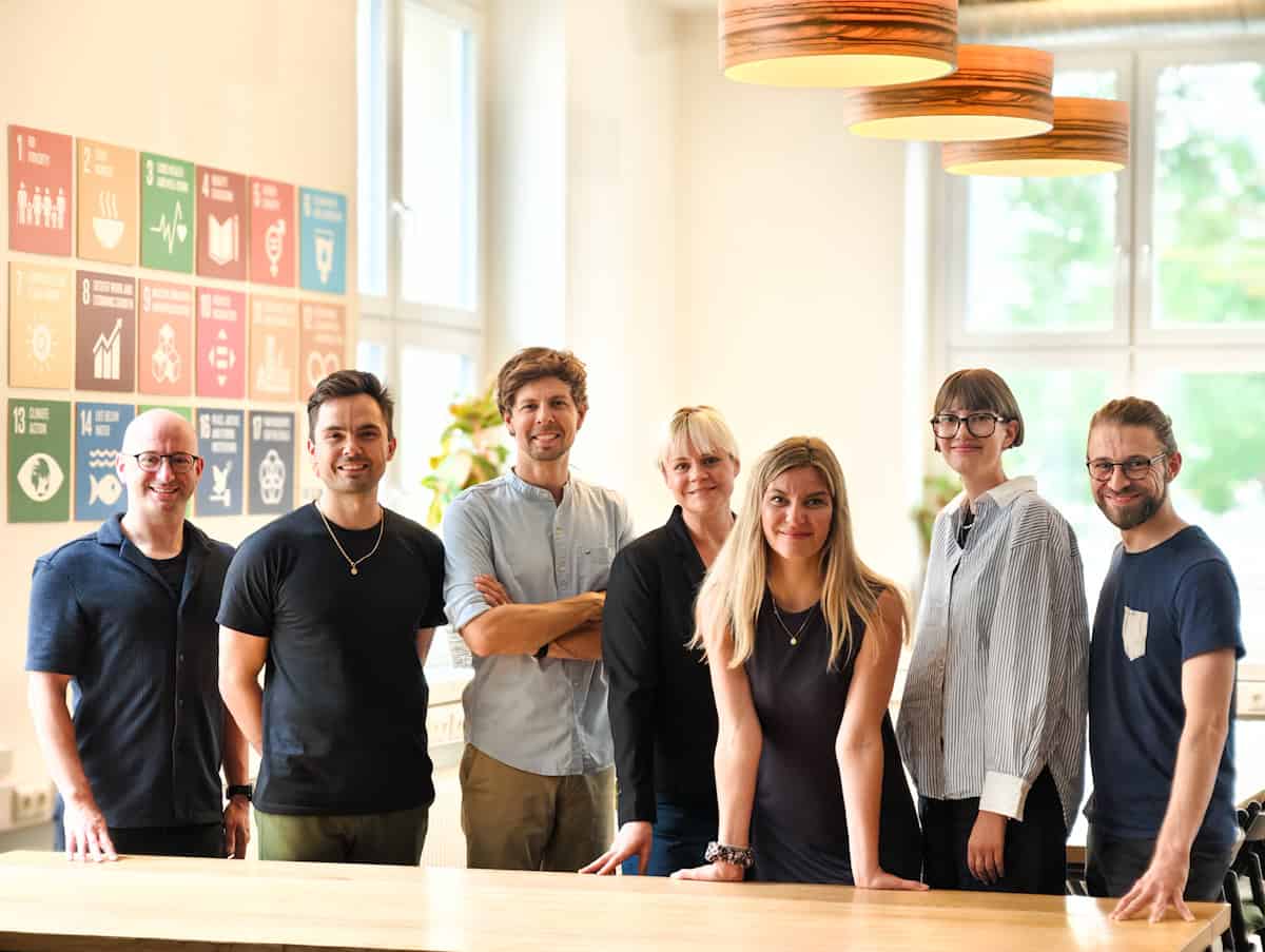 An Tisch stehend von links nach rechts die Teammitglieder Michael Jenkner, Toni Koç, Matthias Damert, Silke Nyvlt, Franziska Kramer, Emma Deeken und Steve Grundig. Sie stehen gemeinsam im Co-Working Space Impact Hub Dresden, dem Firmensitz von plant values