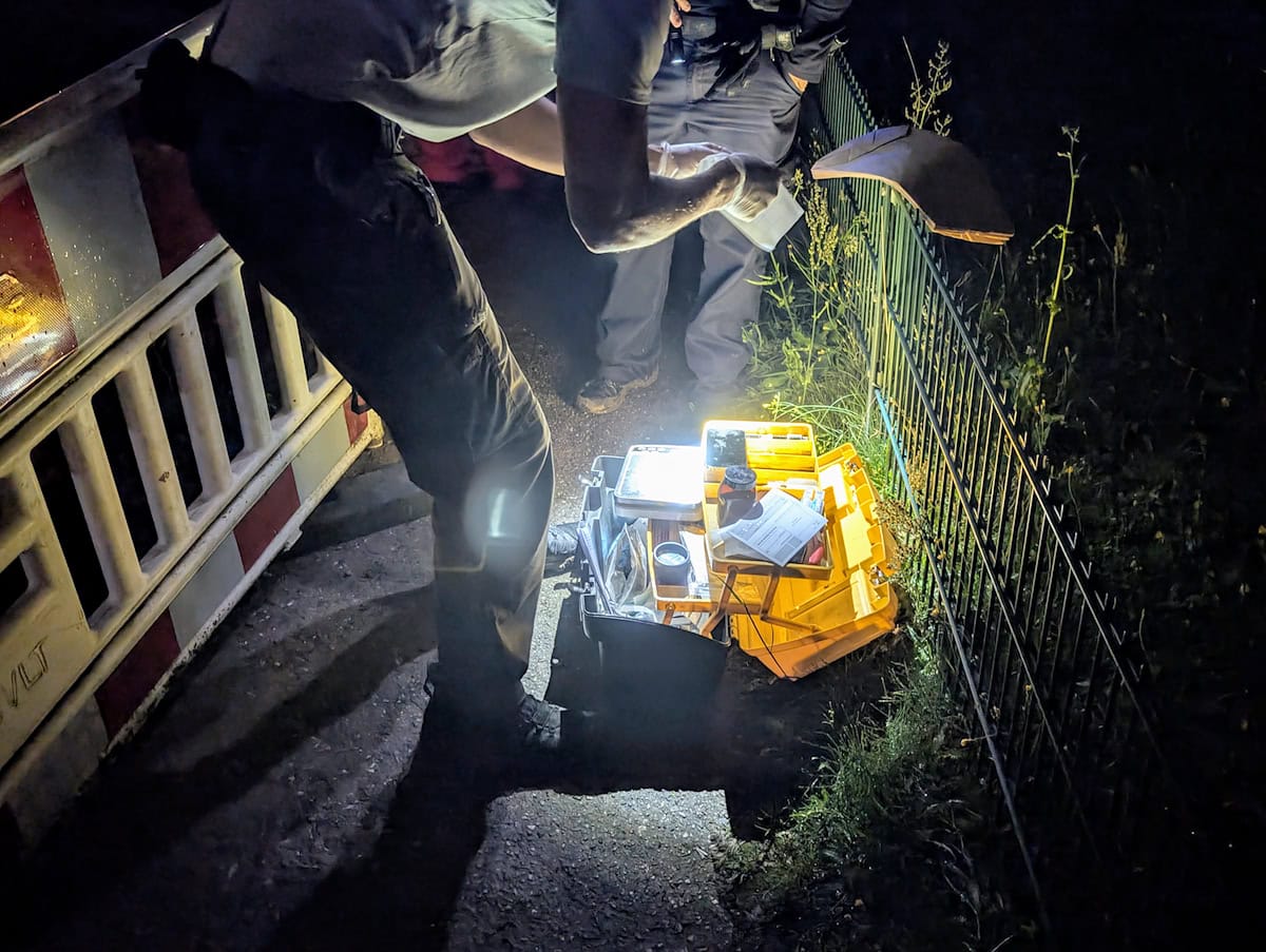 Polizist bei nächtlicher Spurensicherung mit Ausrüstungskoffer und Dokumenten neben einem Zaun. Polizist bei nächtlicher Spurensicherung mit Ausrüstungskoffer und Dokumenten neben einem Zaun