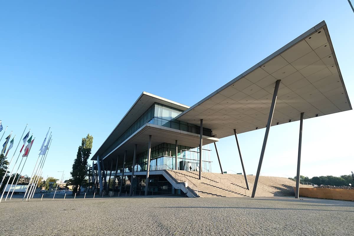 Modernes Gebäude mit großer, schräg geneigter Dachfläche und breiter Treppe im Vordergrund bei klarem Himmel Modernes Gebäude mit großer, schräg geneigter Dachfläche und breiter Treppe im Vordergrund bei klarem Himmel