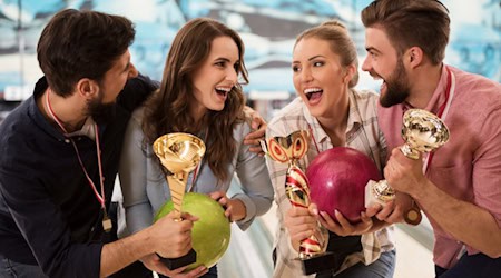 Vier Erwachsene feiern in einer Bowlinghalle mit Pokalen und Bowlingkugeln einen gemeinsamen Sieg.