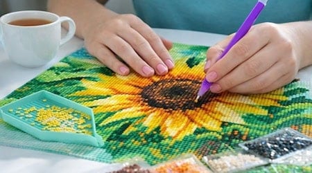 Hände platzieren mit einem violetten Stift kleine Steinchen auf ein Sonnenblumen-Motiv beim Diamond Painting; daneben liegen Sortierschale, Beutel mit Steinen und eine Tasse Tee.