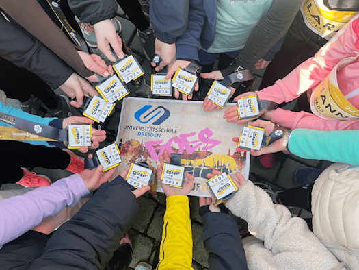 Um ein Schild mit der Aufschrift "Universitätsschule Dresden goes novaled Citylauf Dresden 2026" halten 12 Kinderhände eine Teilnehmerkarte für den Citylauf. Auf dem Schild sind die Silhouette der Stadt Dresden, ein Laufschuhabdruck und das Logo der Universitätsgemeinschaftsschule abgebildet.
