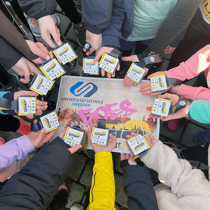 Um ein Schild mit der Aufschrift "Universitätsschule Dresden goes novaled Citylauf Dresden 2026" halten 12 Kinderhände eine Teilnehmerkarte für den Citylauf. Auf dem Schild sind die Silhouette der Stadt Dresden, ein Laufschuhabdruck und das Logo der Universitätsgemeinschaftsschule abgebildet.