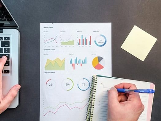 Eine Person analysiert Diagramme und Notizen auf einem Blatt Papier, während sie am Laptop arbeitet – vermutlich zur Auswertung von Vergleichsdaten oder Marktanalysen.