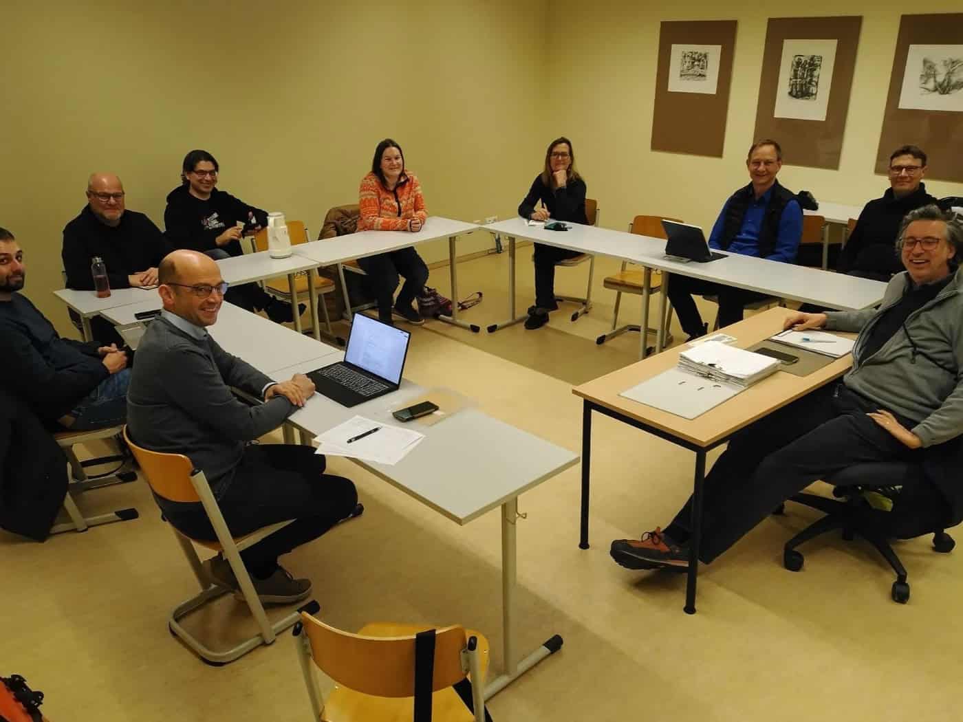 Eine Gruppe sitzt in einem Seminarraum vor Unterlagen und schaut lächelnd in die Kamera