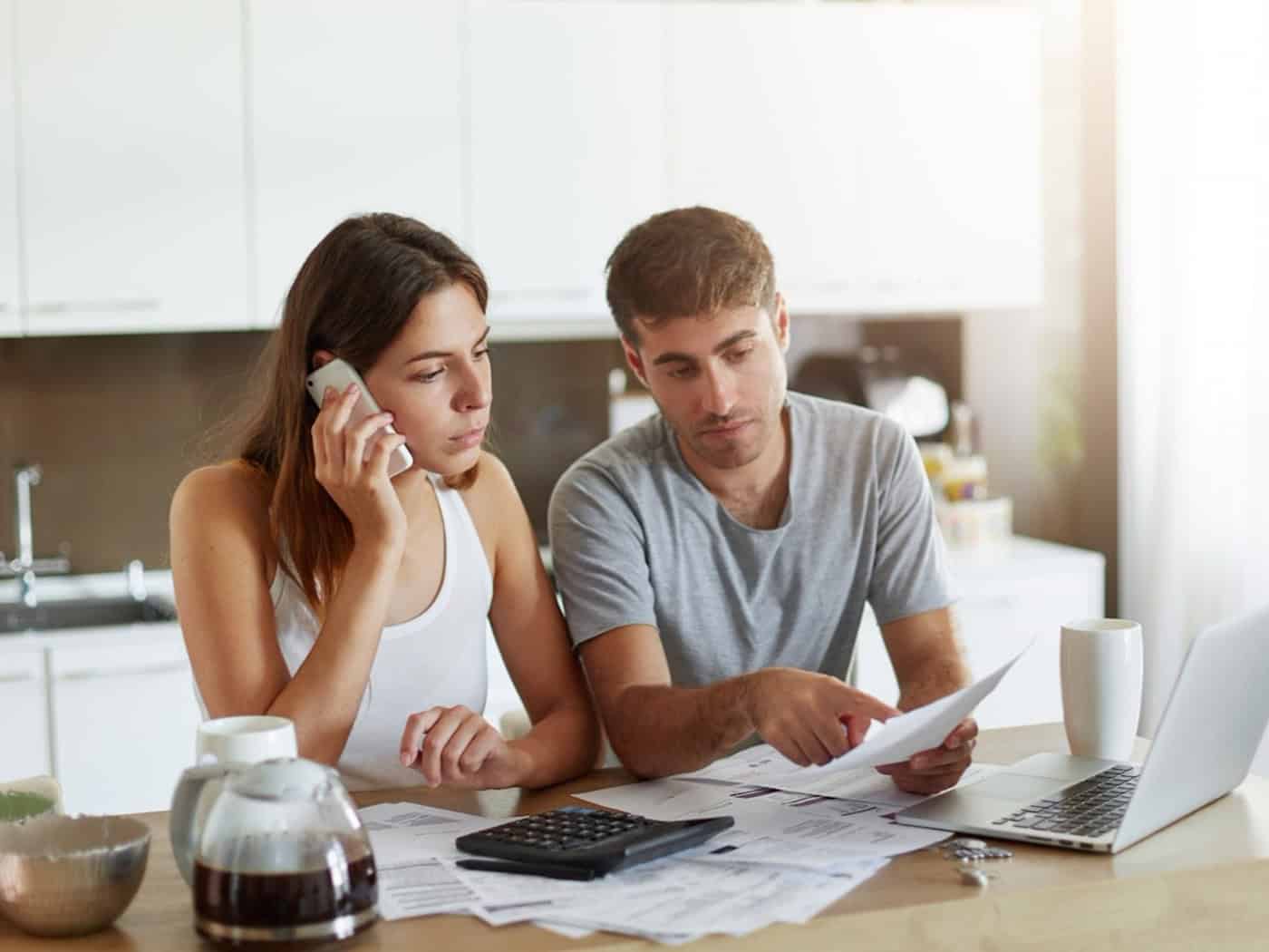 Paar bespricht Finanzunterlagen am Küchentisch – sie ruft einen Finanzberater an, während er Dokumente prüft.