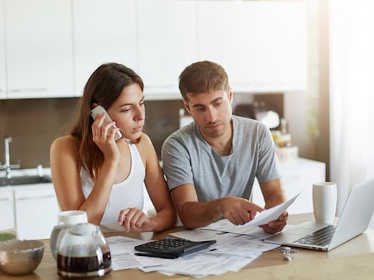 Paar bespricht Finanzunterlagen am Küchentisch – sie ruft einen Finanzberater an, während er Dokumente prüft.