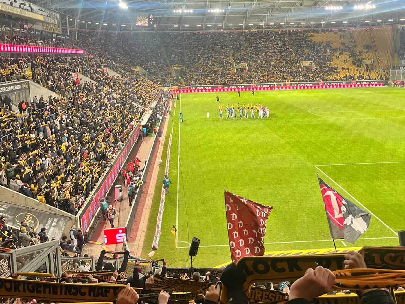 Blick ins ausverkaufte Rudolf-Harbig-Stadion bei einem Abendspiel von Dynamo Dresden, mit dicht gefüllten Rängen in Gelb und Schwarz und beiden Teams zur Platzbegehung im Mittelkreis.
