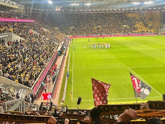 Blick ins ausverkaufte Rudolf-Harbig-Stadion bei einem Abendspiel von Dynamo Dresden, mit dicht gefüllten Rängen in Gelb und Schwarz und beiden Teams zur Platzbegehung im Mittelkreis.