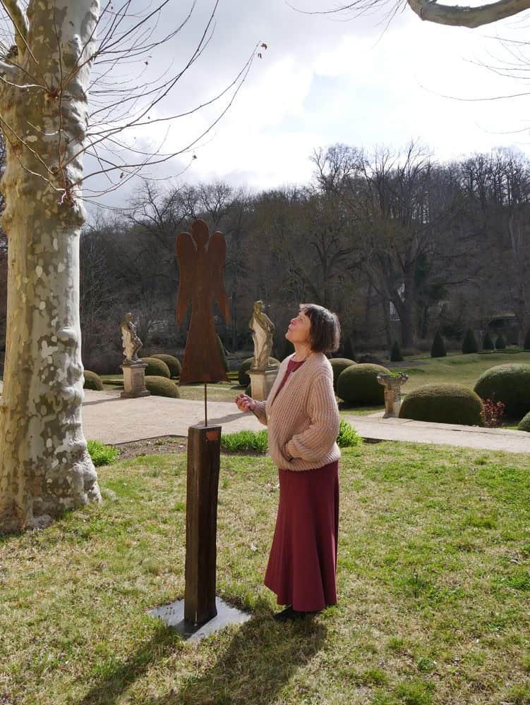 Foto: eine ältere Frau betrachtet eine Skulptur eines stilisierten Engels aus Metall, in einer Parklandschaft