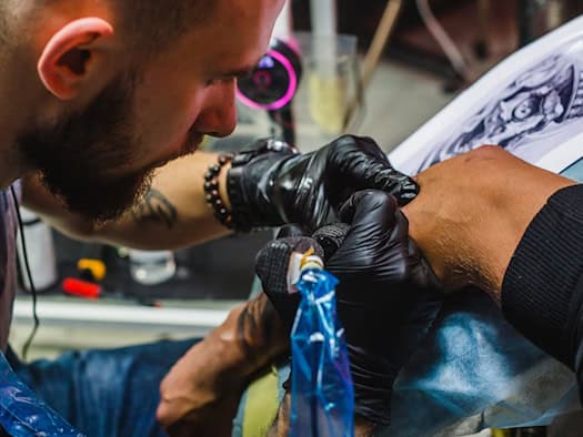 Tätowierer bei der Arbeit in einem Tattoo-Studio, trägt schwarze Handschuhe und arbeitet konzentriert mit einer Tattoo-Maschine am Arm eines Kunden.