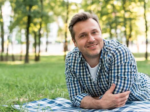 Lächelnder Mann um die 50 in entspannter Haltung auf einer Decke im Grünen – Symbol für Lebensqualität und Selbstbewusstsein auch im höheren Alter