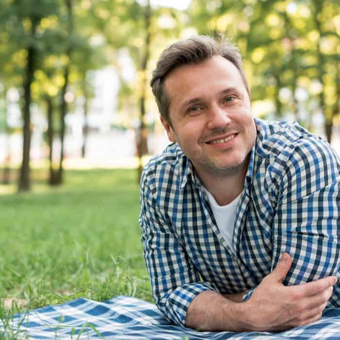 Lächelnder Mann um die 50 in entspannter Haltung auf einer Decke im Grünen – Symbol für Lebensqualität und Selbstbewusstsein auch im höheren Alter