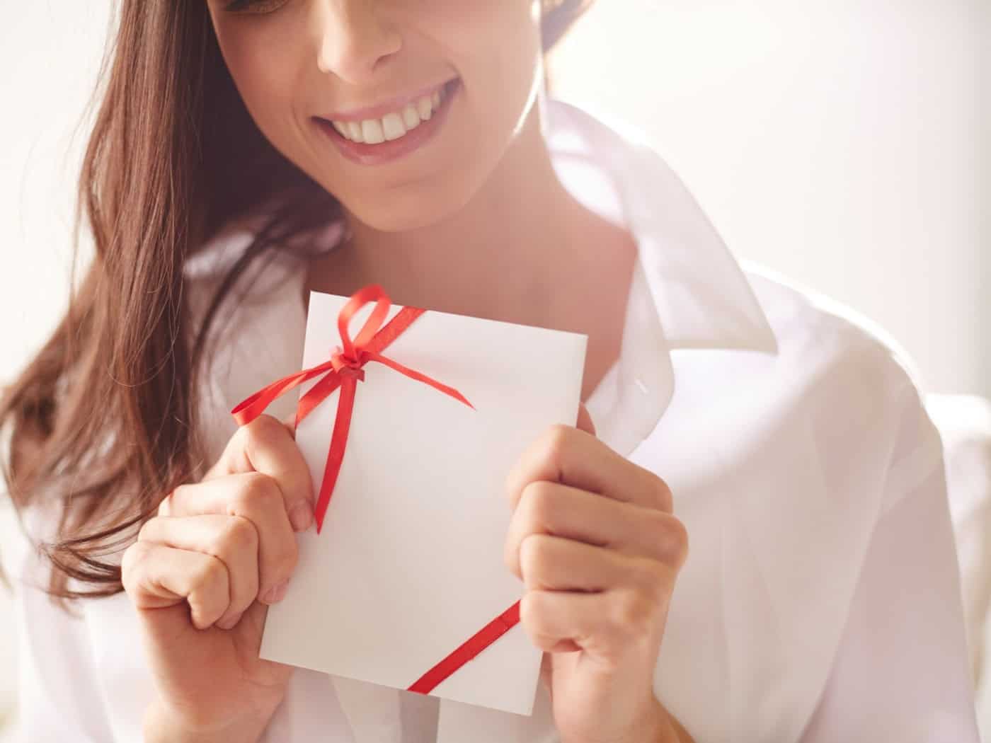 Eine lächelnde Frau hält einen weißen Geschenkgutschein mit roter Schleife in den Händen, symbolisch für die Freude über ein Erlebnisgeschenk.