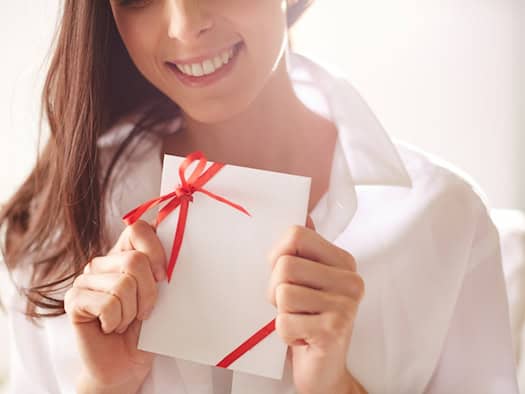 Eine lächelnde Frau hält einen weißen Geschenkgutschein mit roter Schleife in den Händen, symbolisch für die Freude über ein Erlebnisgeschenk.