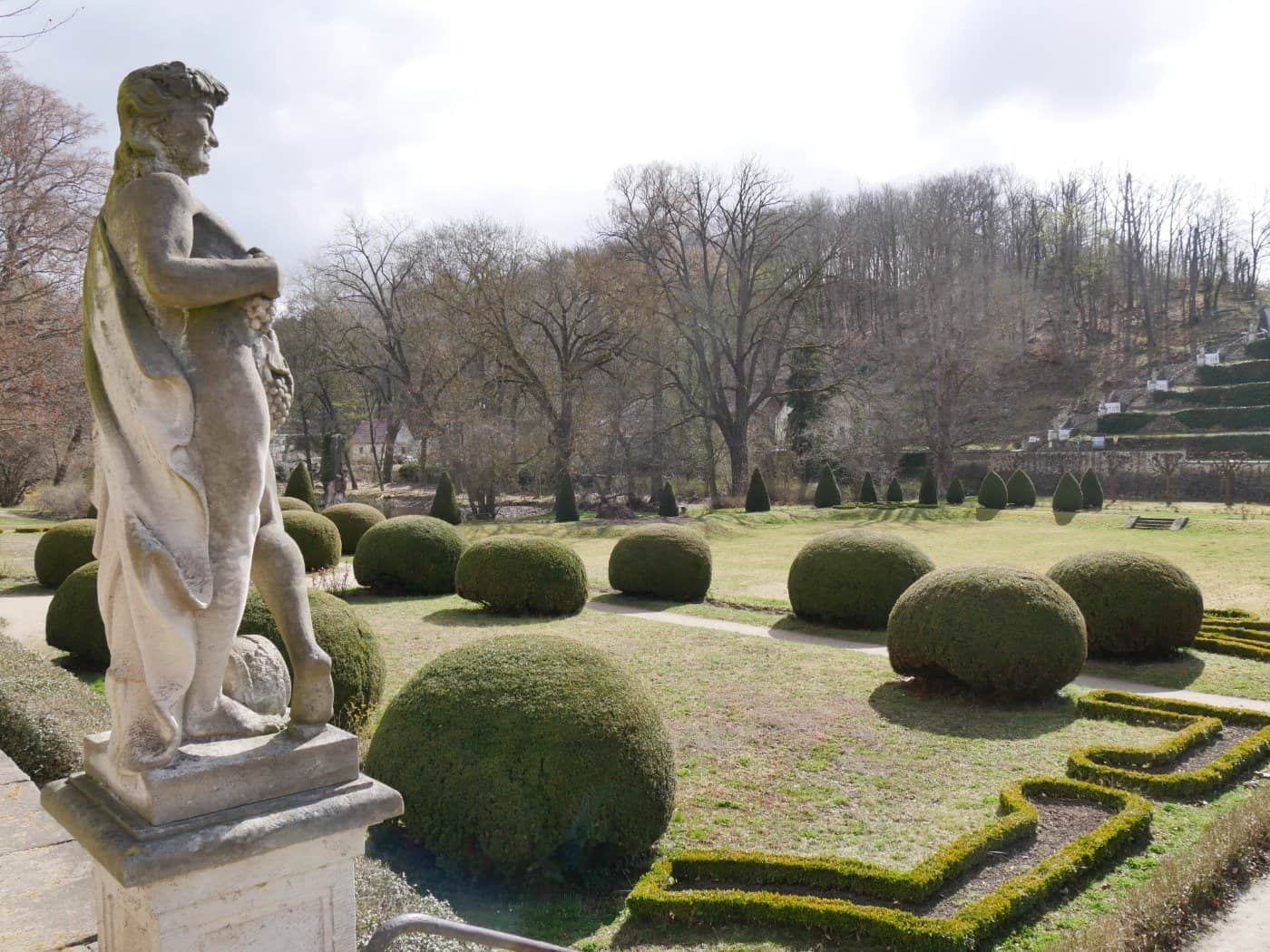 Foto: Blick auf eine gepflegte, winterliche Parkanlage mit Wiesen und Büschen, im Vordergrund eine Steinstatue, im Hintergrund ein bewaldeter Hügel