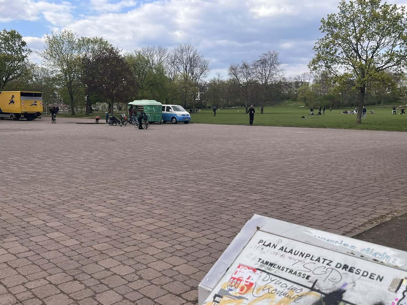 Ein Blick über den gepflasterten Randbereich des Alaunparks in Dresden. Im Vordergrund ist eine Infotafel mit dem Lageplan des Parks zu sehen, im Hintergrund stehen Lieferwagen für den Marktabbau.