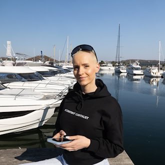 Anike Ekina, Content-Creatorin und Unternehmerin, hält ein Smartphone in der Hand und steht vor einem Yachthafen.