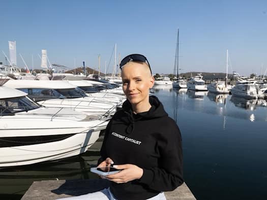 Anike Ekina, Content-Creatorin und Unternehmerin, hält ein Smartphone in der Hand und steht vor einem Yachthafen.