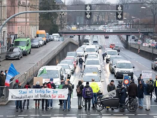 Archivbild einer Klimaschutz-Blockade in Dresden: Aktivisten von Extinction Rebellion stehen im Januar 2026 mit einem Banner auf der Kreuzung Stauffenbergallee und Königsbrücker Straße. Das Banner trägt die Aufschrift „Stau ist lästig für Sie, Klimakollaps ist tödlich für alle“. Im Hintergrund staut