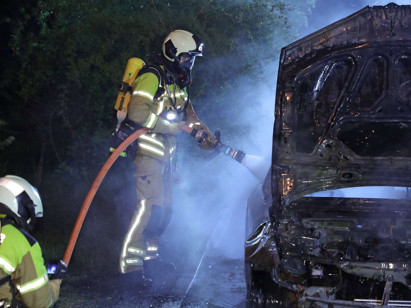 Zwei Feuerwehrleute der Feuerwehr Dresden im nächtlichen Einsatz bei einem Autobrand. Ein Feuerwehrmann unter Atemschutz richtet ein Strahlrohr auf den ausgebrannten Motorraum eines Pkw, dessen Motorhaube offen steht. Dichter weißer Rauch und Wasserdampf steigen auf. Im Vordergrund ist ein weiterer 