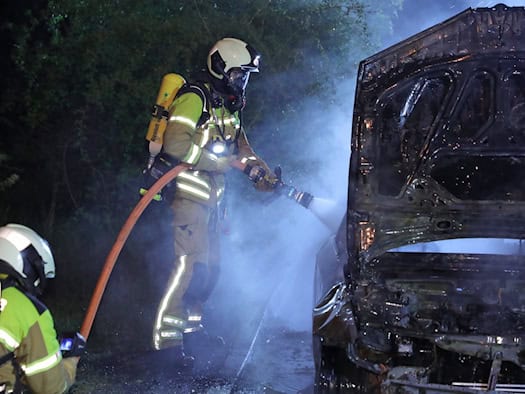 Zwei Feuerwehrleute der Feuerwehr Dresden im nächtlichen Einsatz bei einem Autobrand. Ein Feuerwehrmann unter Atemschutz richtet ein Strahlrohr auf den ausgebrannten Motorraum eines Pkw, dessen Motorhaube offen steht. Dichter weißer Rauch und Wasserdampf steigen auf. Im Vordergrund ist ein weiterer 