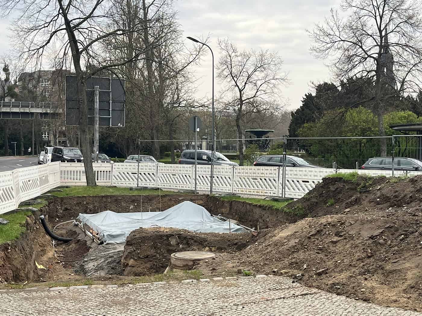 Baustelle für eine neue öffentliche Toilette am Albertplatz in Dresden; ausgehobene Grube mit Fundamentvorbereitung und Absperrzäunen vor Parkhintergrund.