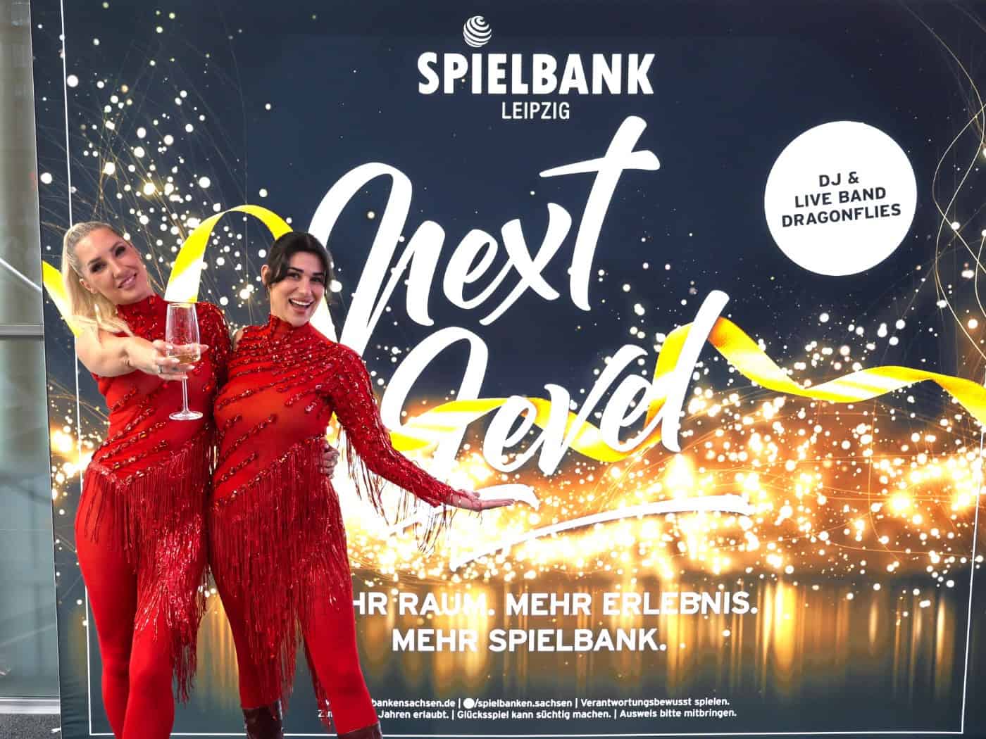 Zwei lächelnde Damen in glitzernden roten Abendoutfits posieren vor dem „Next Level“-Plakat der Spielbank Leipzig und halten ein Sektglas in der Hand.