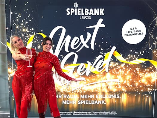 Zwei lächelnde Damen in glitzernden roten Abendoutfits posieren vor dem „Next Level“-Plakat der Spielbank Leipzig und halten ein Sektglas in der Hand.