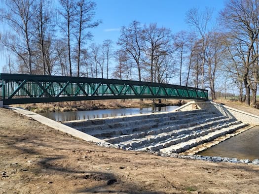 Foto: schmale, lange Fußgängerbrücke über ein Wehr eines Flusses, im Hintergrund Bäume ohne Blätter vor blauem Himmel