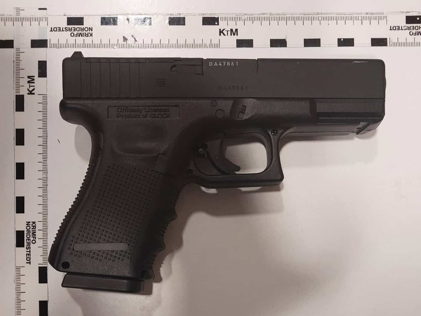 Eine schwarze, offiziell lizenzierte Glock-Nachbildung (Diabolo-Waffe) mit eingelegtem Magazin liegt auf einer weißen Unterlage mit polizeilichen Maßstäben der Kriminaltechnik (KTM).