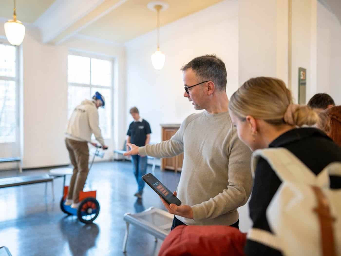 Ein Dozent der HTW Dresden erklärt einer Gruppe interessierter junger Leute eine Anwendung auf einem Tablet-PC. Im Hintergrund balanciert eine Person in einer hellen Halle auf einem zweirädrigen Elektro-Gefährt (Segway-Typ), was die praxisnahe Forschung im Bereich Mobilität verdeutlicht.