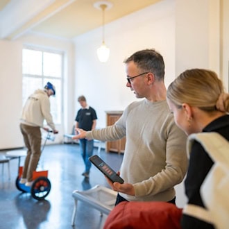 Ein Dozent der HTW Dresden erklärt einer Gruppe interessierter junger Leute eine Anwendung auf einem Tablet-PC. Im Hintergrund balanciert eine Person in einer hellen Halle auf einem zweirädrigen Elektro-Gefährt (Segway-Typ), was die praxisnahe Forschung im Bereich Mobilität verdeutlicht.