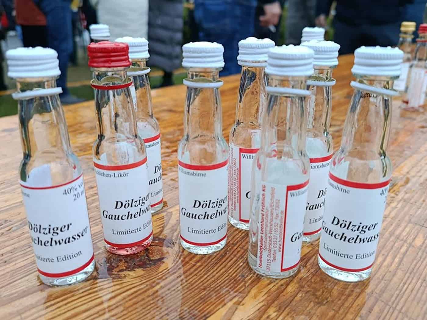 Foto: Nahaufnahme mehrerer leerer Likorfläschchen (20ml) auf einem regennassen Biertisch stehend, im Hintergrund stehen Menschen