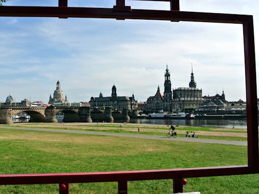 Blick auf die Dresdner Altstadt-Silhouette mit Frauenkirche, Hofkirche und Kunstakademie, eingerahmt durch einen großen, roten Metallrahmen am Elbufer (Canaletto-Blick). Im Vordergrund sind grüne Elbwiesen und ein Radfahrer zu sehen. Das Bild symbolisiert die Diskrepanz zwischen touristischem Glanz 
