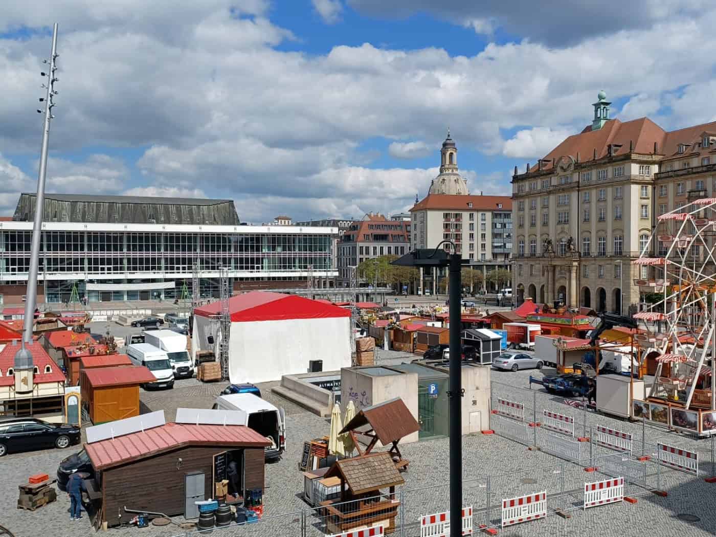 Weitwinkelaufnahme des Dresdner Altmarkts während der Aufbauarbeiten für den Frühjahrsmarkt. Zu sehen sind hölzerne Markthütten, ein Riesenrad, der Kulturpalast und im Hintergrund die Kuppel der Frauenkirche unter einem blau-weißen Frühlingshimmel.