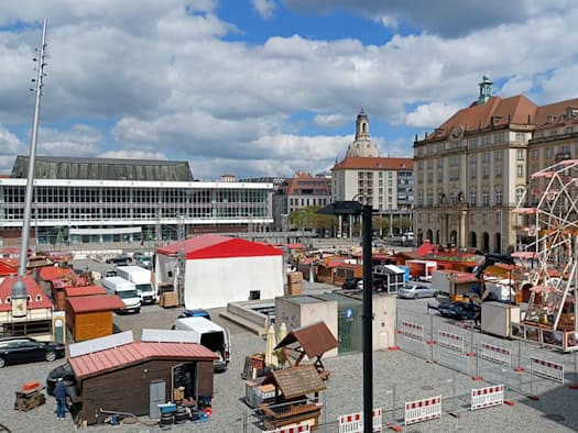 Weitwinkelaufnahme des Dresdner Altmarkts während der Aufbauarbeiten für den Frühjahrsmarkt. Zu sehen sind hölzerne Markthütten, ein Riesenrad, der Kulturpalast und im Hintergrund die Kuppel der Frauenkirche unter einem blau-weißen Frühlingshimmel.