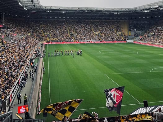 Blick aus der Fanperspektive auf das Spielfeld des Rudolf-Harbig-Stadions in Dresden während des Einlaufens der Mannschaften. Im Vordergrund halten Fans Schals und gelb-schwarze Fahnen der SG Dynamo Dresden hoch; die Zuschauerränge sind voll besetzt unter einem bewölkten Himmel.