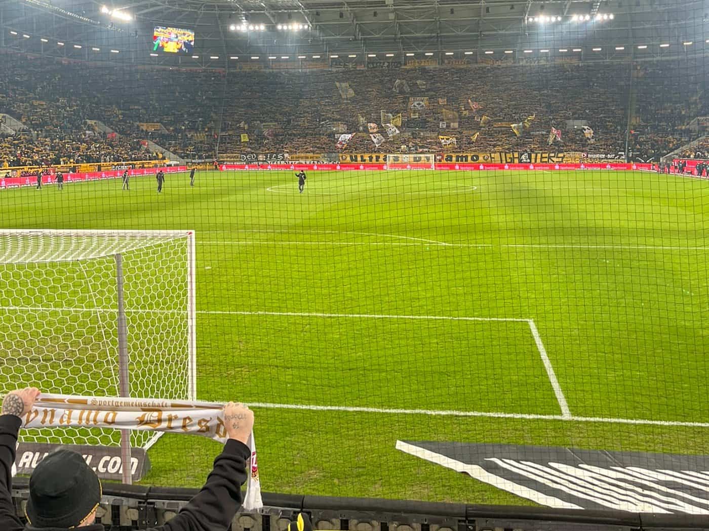 Blick aus der Fanperspektive hinter dem Tor in das vollbesetzte Rudolf-Harbig-Stadion bei einem Abendspiel von Dynamo Dresden. Ein Fan hält einen Vereinsschal hoch, im Hintergrund ist die beleuchtete Tribüne mit zahlreichen gelb-schwarzen Fahnen und Bannern der Ultras zu sehen. Ein Sicherheitsnetz t