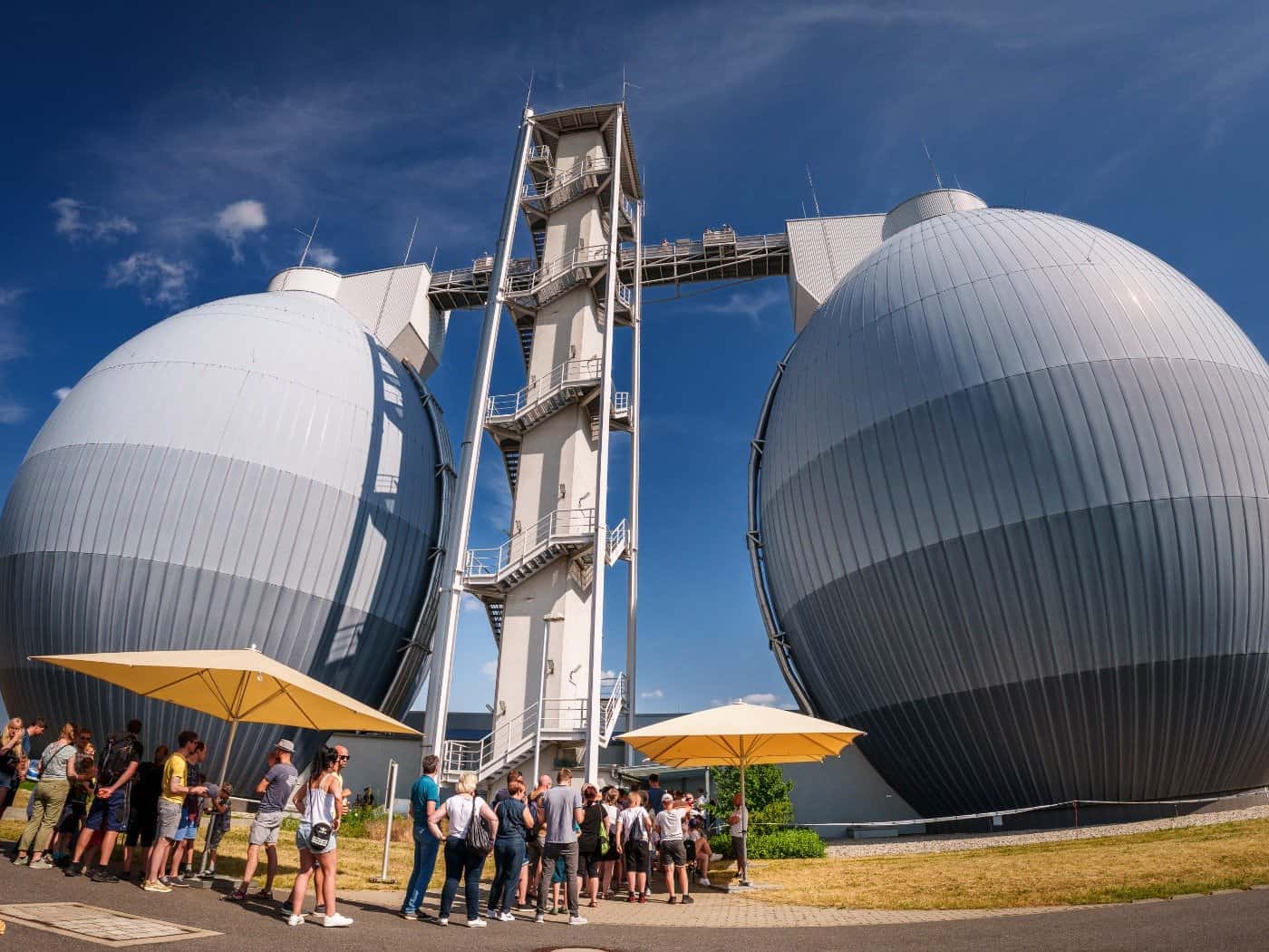 Die zwei großen, eiförmigen Faultürme der Kläranlage Dresden-Kaditz bei sonnigem Wetter. Im Vordergrund stehen Besucher in einer Schlange unter Sonnenschirmen für eine Besichtigung an.