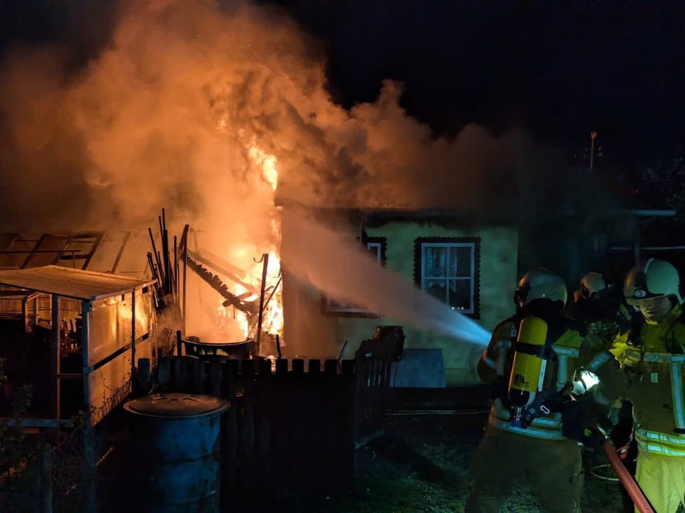 Nachtaufnahme eines Feuerwehreinsatzes in Dresden-Gruna: Mehrere Einsatzkräfte in Schutzkleidung richten kraftvolle Wasserstrahlen auf eine brennende Gartenlaube unter schwarzem Nachthimmel.