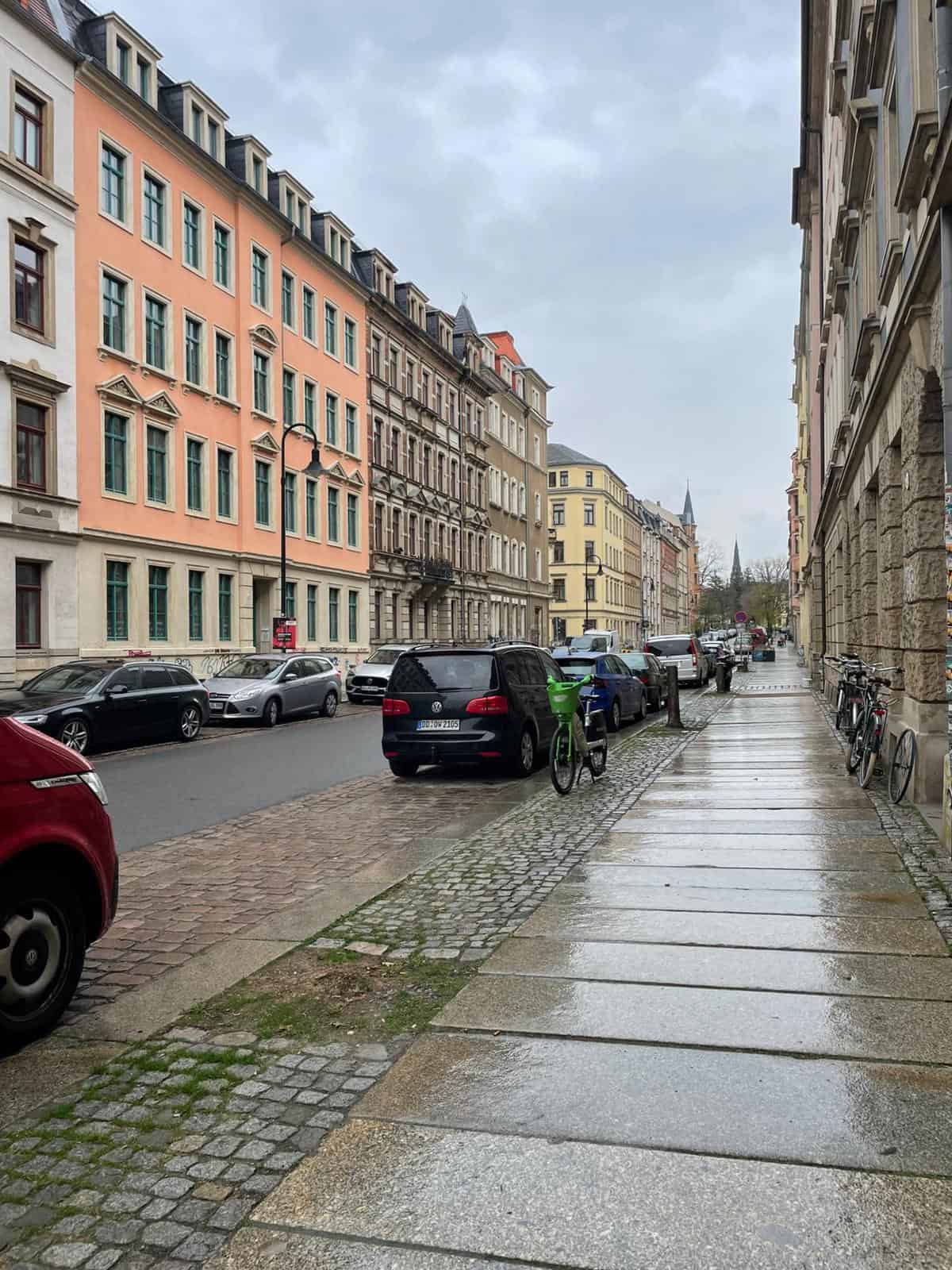 Identischer regennasser Blick die Fichtenstraße in der Dresdner Neustadt hinunter. Geparkte Autos und historische Gründerzeithäuser unter grauem Himmel sind sichtbar. Auf dem Gehweg ist kein Baum und kein markanter Stumpf im unmittelbaren Vordergrund zu sehen, was einen baumlosen Zustand zeigt.