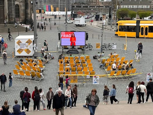 Publikum sitzt in gelben Liegestühlen beim Open-Air-Kino auf dem Dresdner Schloßplatz während des 38. Filmfest Dresden; im Hintergrund eine große LED-Leinwand und eine gelbe Straßenbahn.