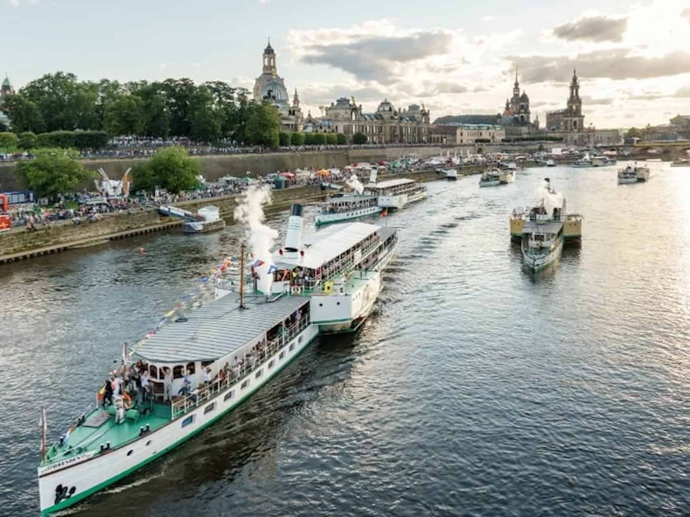 Mehrere historische Raddampfer der Sächsischen Dampfschifffahrt fahren bei sonnigem Wetter auf der Elbe in Dresden, im Hintergrund die Silhouette der Altstadt mit der Frauenkirche.